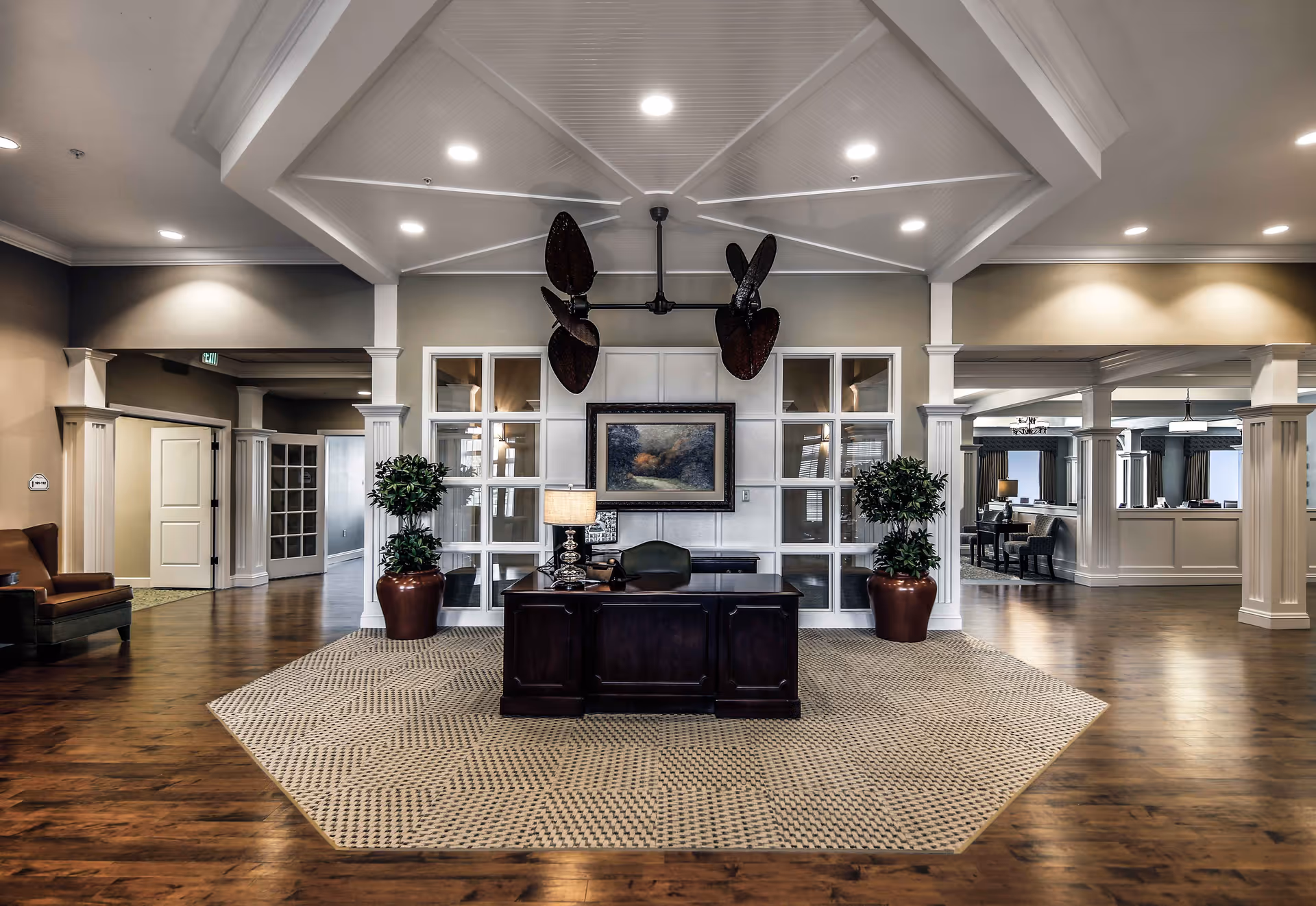 A spacious and elegant reception area with a dark wooden desk centered on a patterned rug. Behind the desk is a wall with large windows and a framed landscape painting. Two large potted plants flank the desk. The ceiling features recessed lighting and a decorative fan with large blades. The floor is wooden, and there are open doorways leading to other rooms.