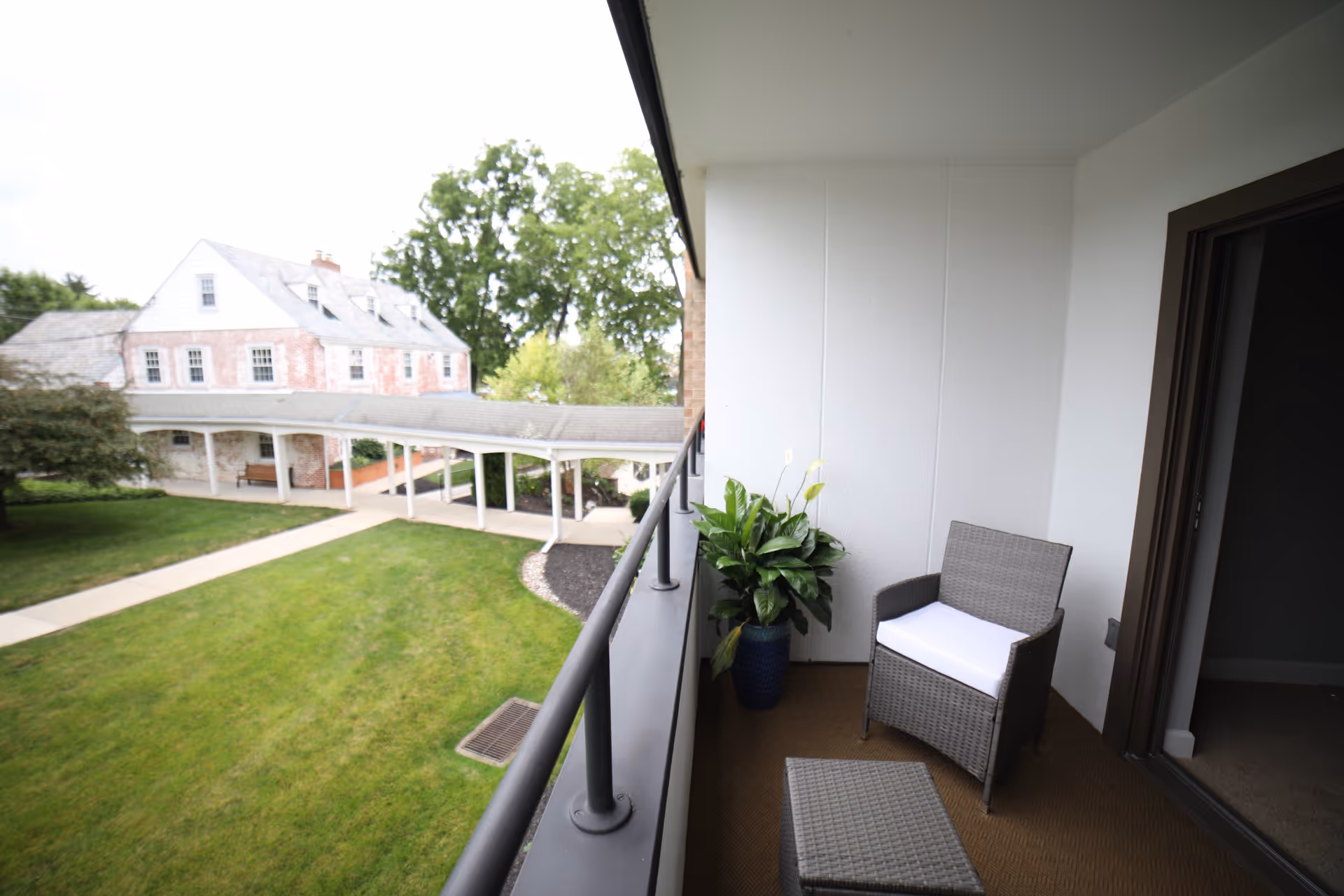 A small balcony with a wicker chair with a white cushion, a matching ottoman, and a potted plant. The balcony overlooks a green lawn with a paved walkway and a large brick building with white trim in the background.