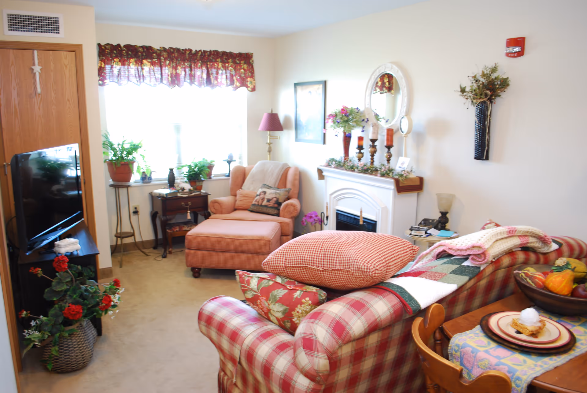 A cozy living room with a red and white plaid sofa adorned with pillows and a quilt. There is a matching armchair with an ottoman near a window with floral curtains. A small wooden side table holds plants and decorative items. A white fireplace mantel is decorated with candles, flowers, and a round mirror above it. A wooden dining table with a colorful tablecloth and a plate with a dessert is partially visible. The room has beige walls and carpeted floor.