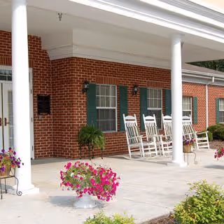 Front porch of a brick building with white columns, several white rocking chairs, and flower pots with pink flowers.