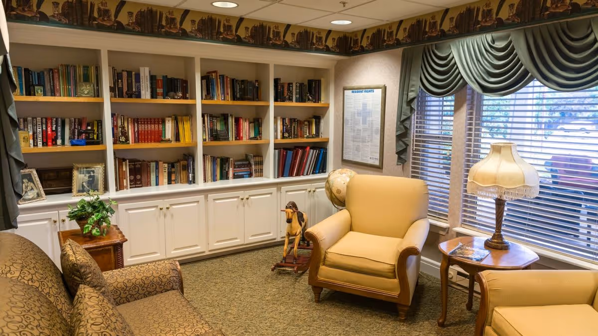 A cozy reading room with built-in white bookshelves filled with books, a beige armchair, a patterned sofa, a wooden side table with a lamp, a rocking horse, and large windows with green valance curtains letting in natural light.