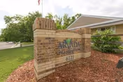 Brick entrance sign reading 'Arvada Care And Rehabilitation Center' with the address '6121' in landscaped grounds and part of the facility building and flagpole visible behind it.