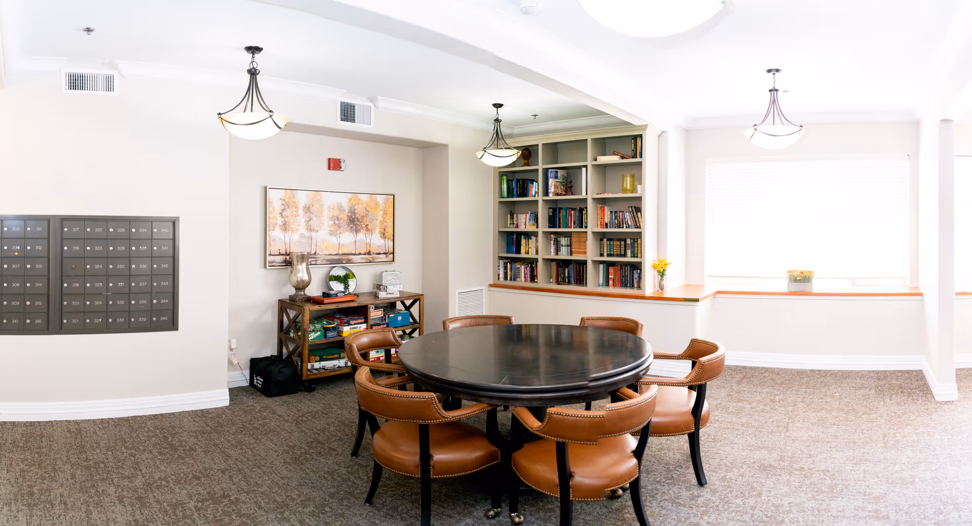 Bright communal interior with a round table surrounded by leather chairs, built-in bookshelves, mailboxes on the wall, and window seating.