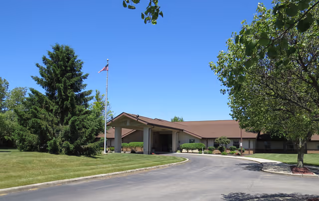 Exterior view of a single-story building with a brown roof and beige walls, surrounded by green trees and a well-maintained lawn. An American flag is flying on a flagpole near the entrance, and a curved driveway leads up to the covered entrance of the building under a clear blue sky.