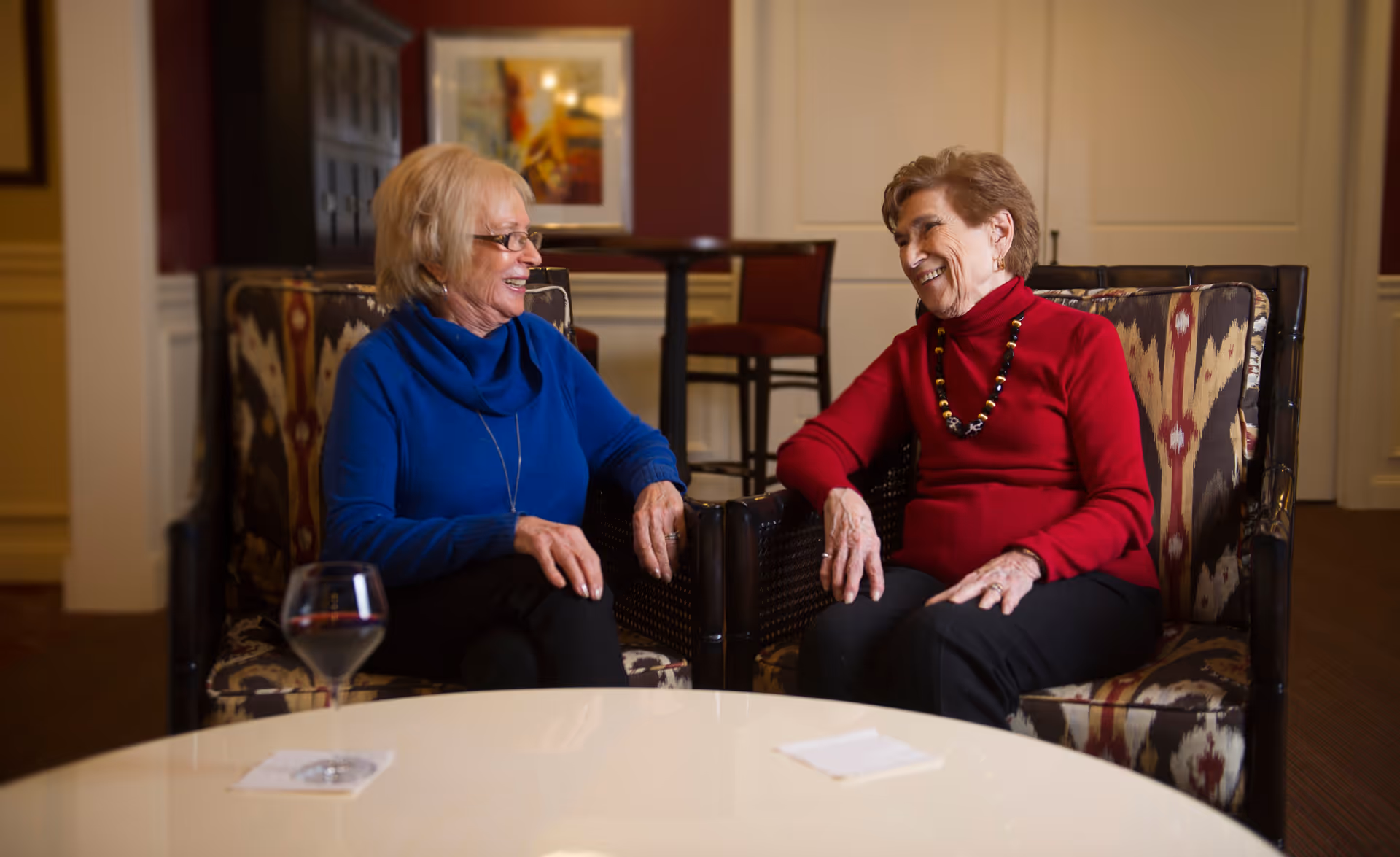 Two elderly women sitting in patterned armchairs facing each other and smiling in a cozy indoor lounge area. A glass of red wine is on a white table in the foreground, and there are chairs and a framed picture in the background.