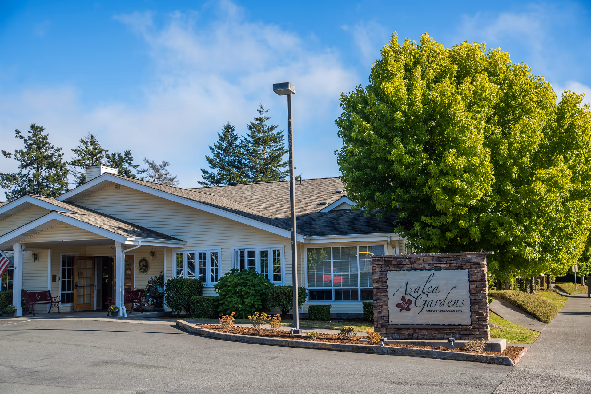 Exterior view of Azalea Gardens Senior Living facility showing a single-story building with a covered entrance, surrounded by greenery and a large tree. A stone sign with the facility name is visible near the sidewalk.