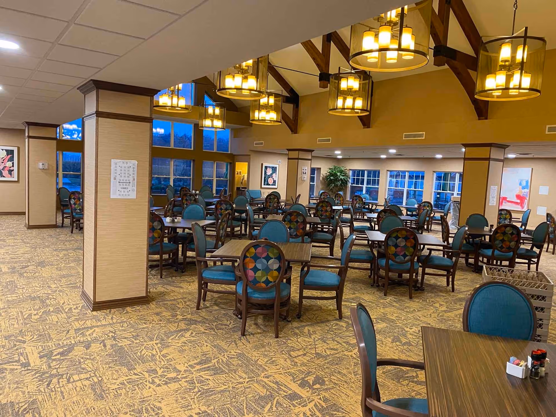 A spacious dining room in a retirement community with multiple wooden tables and chairs featuring colorful patterned backs and teal cushions. The room has large windows, warm lighting from hanging chandeliers, and beige walls with decorative columns.