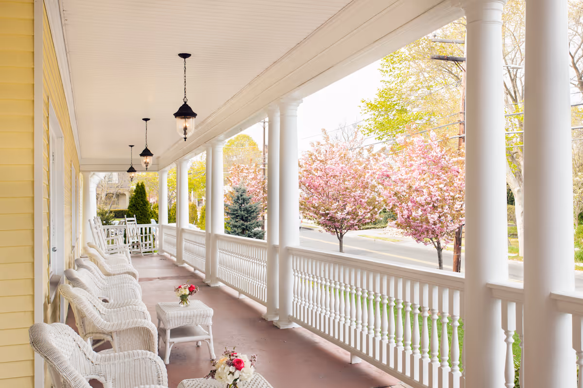 A spacious covered porch with white wicker chairs and tables, decorated with small flower arrangements. The porch has white columns and railings, hanging lantern-style lights, and overlooks a street lined with blooming pink and green trees.