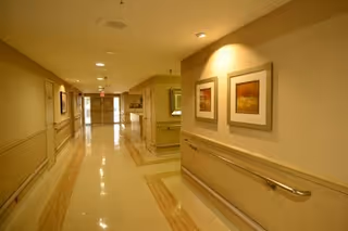 Well-lit hallway inside a care facility with handrails, framed artwork, and doors along the corridor.