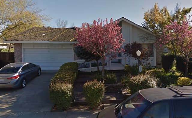 Front exterior view of a single-story house with a two-car garage, a driveway with two parked cars, and a garden with blooming pink trees and green shrubs.