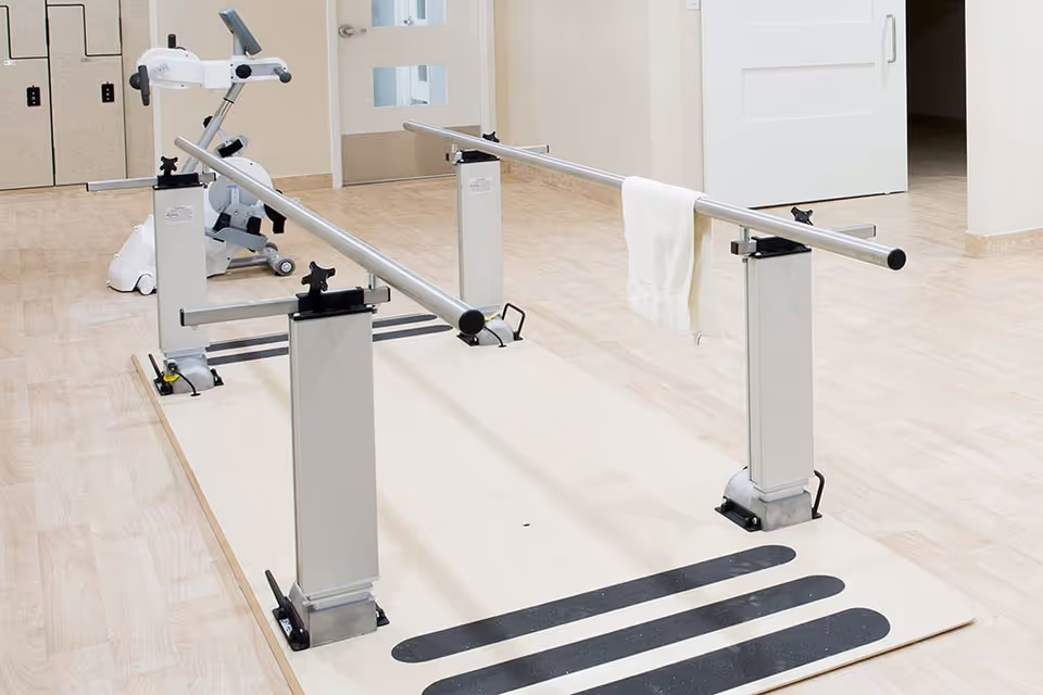 Physical therapy parallel bars setup on a light wood floor with a white towel hanging on one bar and an exercise bike in the background in a bright room with beige walls and doors.