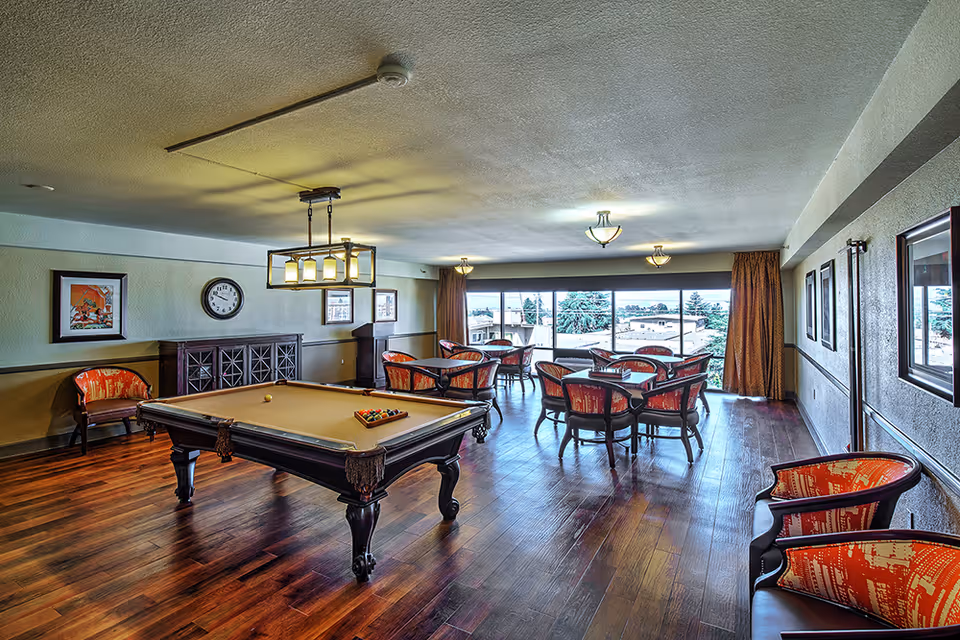 A spacious senior living common room with a pool table in the center, surrounded by several seating areas with tables and chairs. The room has wooden flooring, framed artwork on the walls, a clock, and large windows letting in natural light with a view of trees and buildings outside.