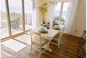 Dining area with a white table set for four, including plates, bowls, and glasses. The room has large sliding glass doors leading to a balcony with outdoor seating and a view of the water. There is a potted plant in the corner and wooden flooring throughout.