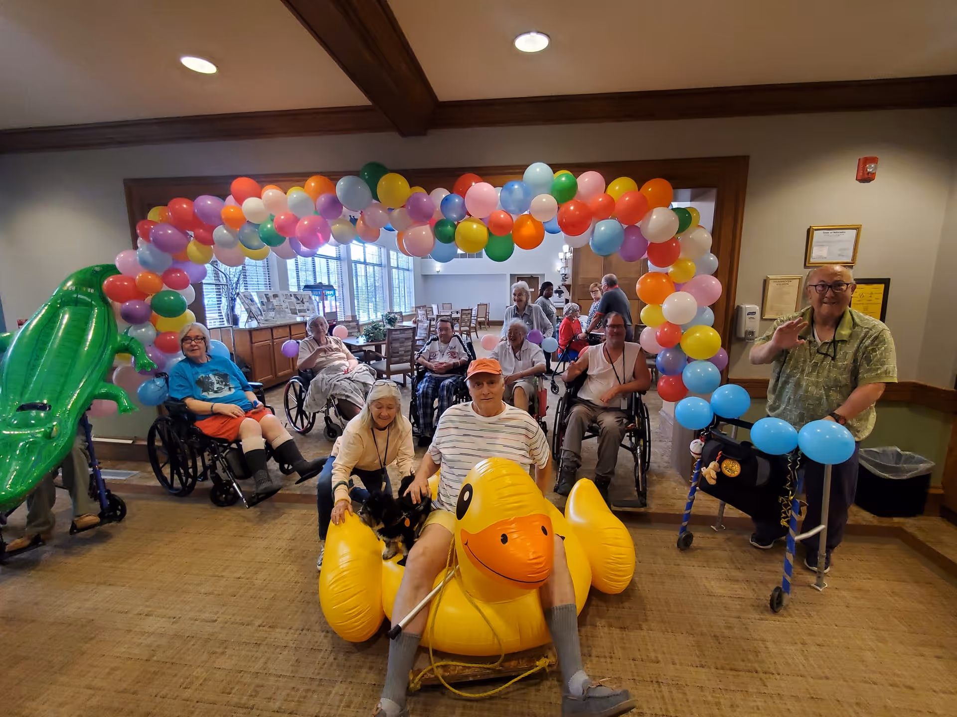 A group of senior residents in a communal indoor space posing under a colorful balloon arch with an oversized inflatable duck and pool float.
