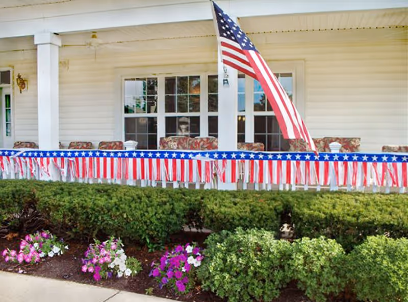 Front porch of a building decorated with American flag-themed bunting and an American flag mounted on a white column. The porch has cushioned chairs and is bordered by green bushes and colorful flowers in the foreground.