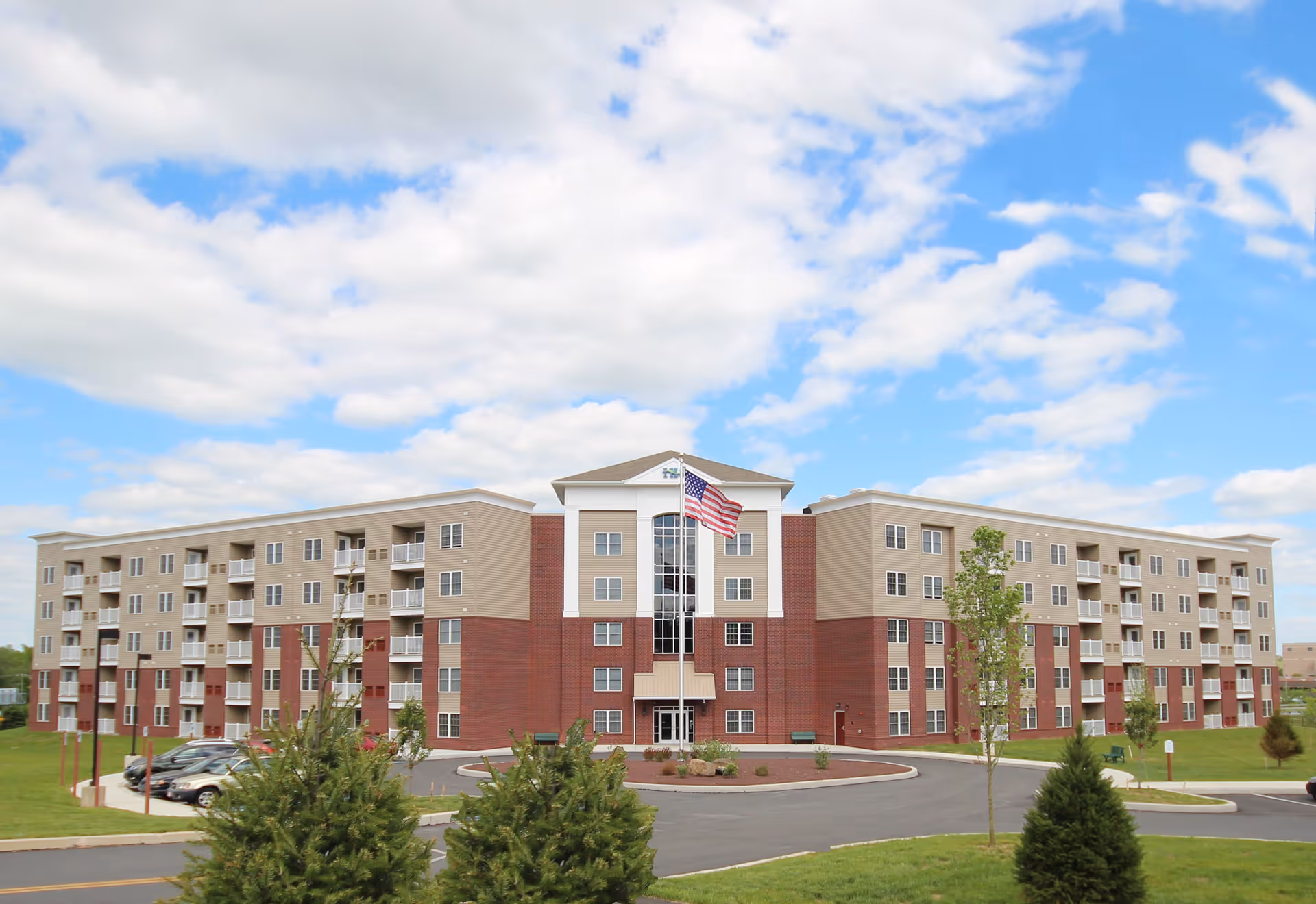 Exterior view of a large, modern four-story apartment building with a combination of red brick and beige siding. The building has multiple balconies and windows, with an American flag flying on a flagpole in front. The surrounding area includes a paved driveway, green lawns, and a few small trees under a partly cloudy blue sky.