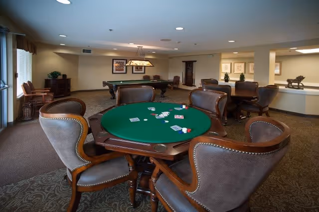 A senior living facility game room with a green poker table surrounded by six cushioned chairs. Playing cards and poker chips are scattered on the table. In the background, there is a pool table with a hanging light fixture above it, additional seating, framed artwork on the walls, and a carpeted floor.