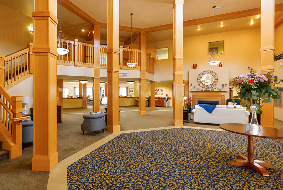 Spacious senior living facility common area with tall wooden pillars, a staircase, carpeted floor, comfortable seating including a white sofa and gray chairs, a fireplace with a decorative mirror above it, and a round wooden table with a vase of flowers.