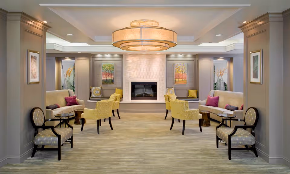 A spacious and elegantly decorated living room area with a central fireplace surrounded by built-in seating. The room features beige and yellow upholstered chairs, sofas with colorful cushions, framed artwork on the walls, and a large circular ceiling light fixture. The walls are painted in neutral tones, and the floor is carpeted.