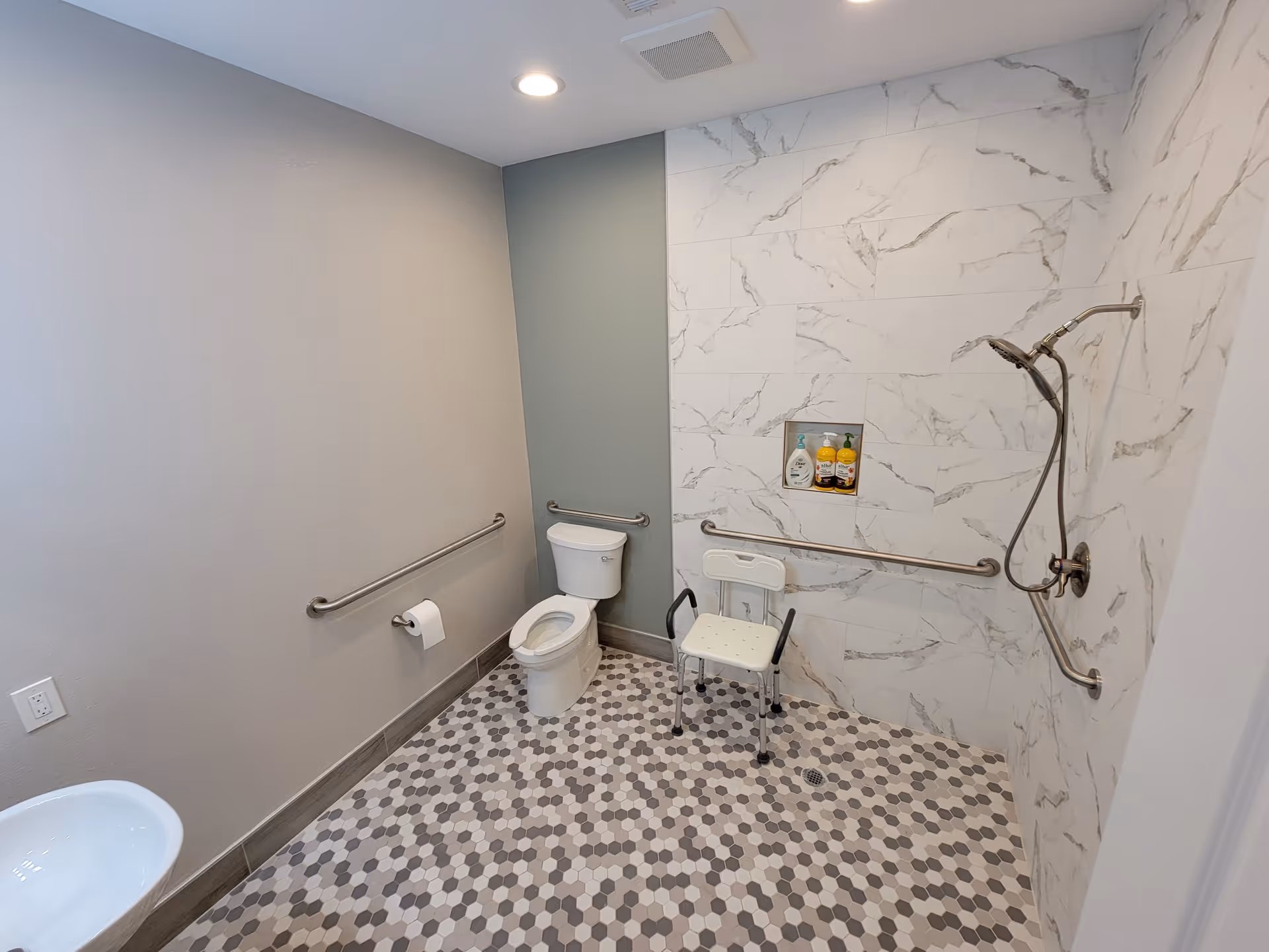 Accessible bathroom with a toilet, a wall-mounted sink, grab bars, a handheld showerhead, a shower chair, and a tiled floor with hexagonal patterns. The shower area has marble-patterned wall tiles and a built-in niche holding toiletries.