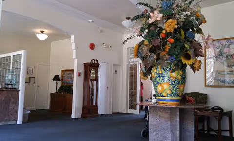 Interior view of a senior living facility lobby area with a large decorative floral arrangement in a colorful vase on a pedestal. The space features carpeted floors, white walls, a grandfather clock, framed artwork, and a water dispenser in the background.