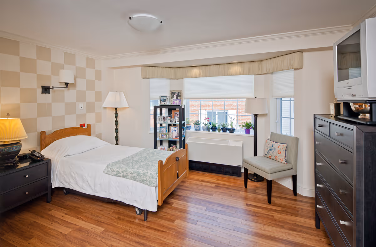 Well-lit bedroom with a single wooden bed, nightstands and lamps, a tall dresser, a chair, and a large window with potted plants.