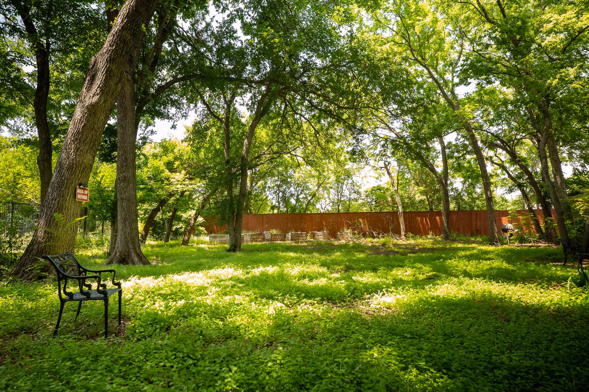 A peaceful outdoor garden area with lush green grass and tall trees providing shade. There are two black metal benches placed on the grass, and a wooden fence in the background. A small sign on one tree reads 'Area of Refuge'.