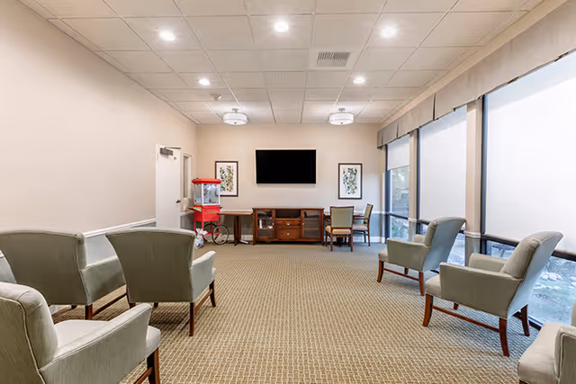 A spacious common area with beige carpet and light-colored walls, featuring several upholstered armchairs arranged facing a wall-mounted flat-screen TV. Below the TV is a wooden cabinet with two framed botanical prints on either side. Large windows with roller shades allow natural light to fill the room. A red popcorn machine is positioned near a door on the left side of the room.