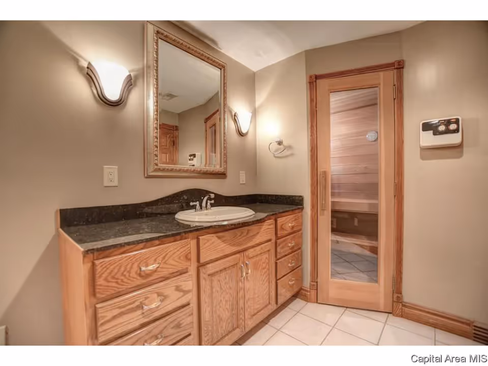 Bathroom vanity with a black granite countertop, a white sink, wooden cabinets, a large framed mirror above the sink, two wall-mounted light fixtures, and a wooden door with a glass panel leading to a sauna room.