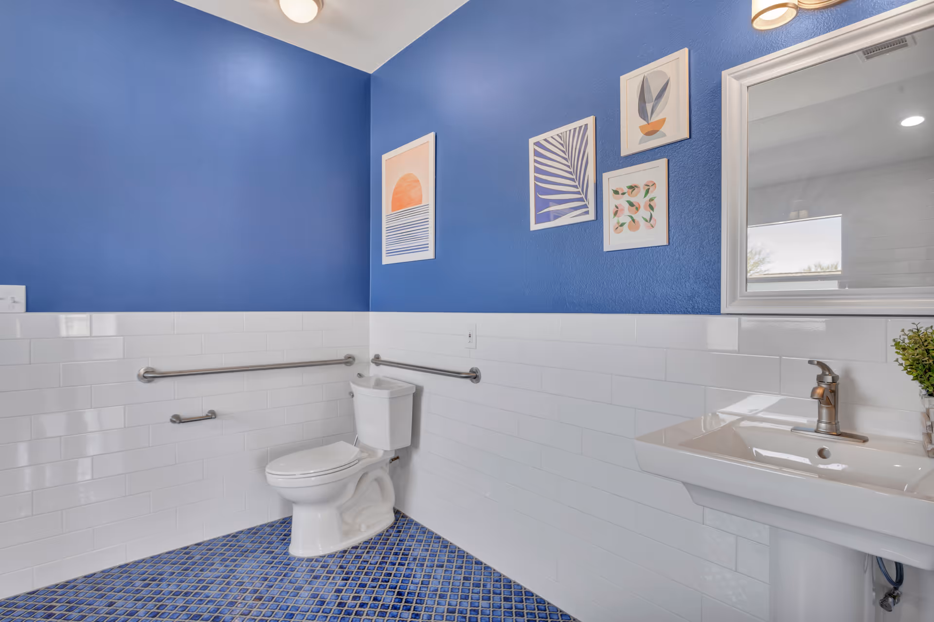 A clean and accessible bathroom with blue tiled floor and blue walls above white subway tiles. The bathroom features a white toilet with grab bars on the walls for support, a white pedestal sink with a modern faucet, a mirror above the sink, and four framed abstract art pieces on the wall.