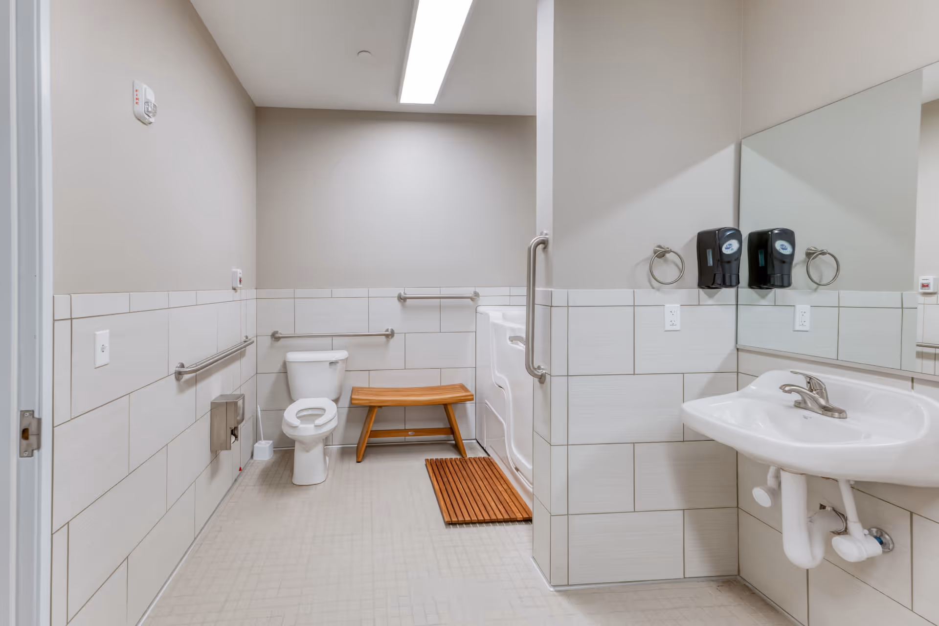 A clean, accessible bathroom with a toilet, a wooden bench, a walk-in shower with a wooden mat, a wall-mounted sink with a faucet, soap dispensers, and grab bars on the walls. The walls are tiled halfway up and painted light gray above, with a large mirror above the sink.