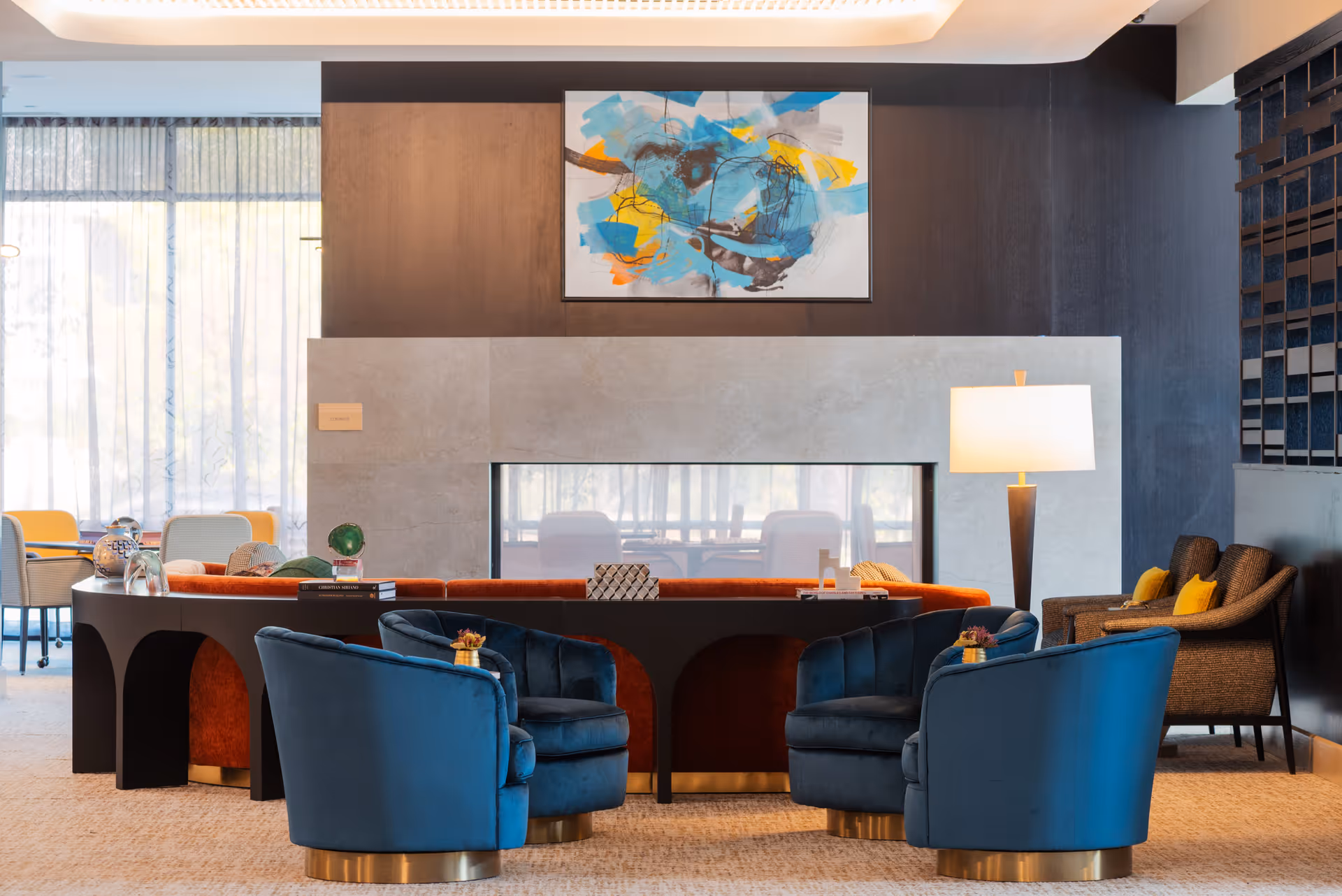 A modern lounge area with four blue velvet armchairs arranged around a black table with decorative items. Behind the table is a long orange sofa facing a double-sided fireplace set in a beige stone wall. Above the fireplace hangs an abstract painting with blue, yellow, and orange colors. To the right, there is a floor lamp and two brown armchairs with yellow cushions. Large windows with sheer curtains allow natural light into the space.