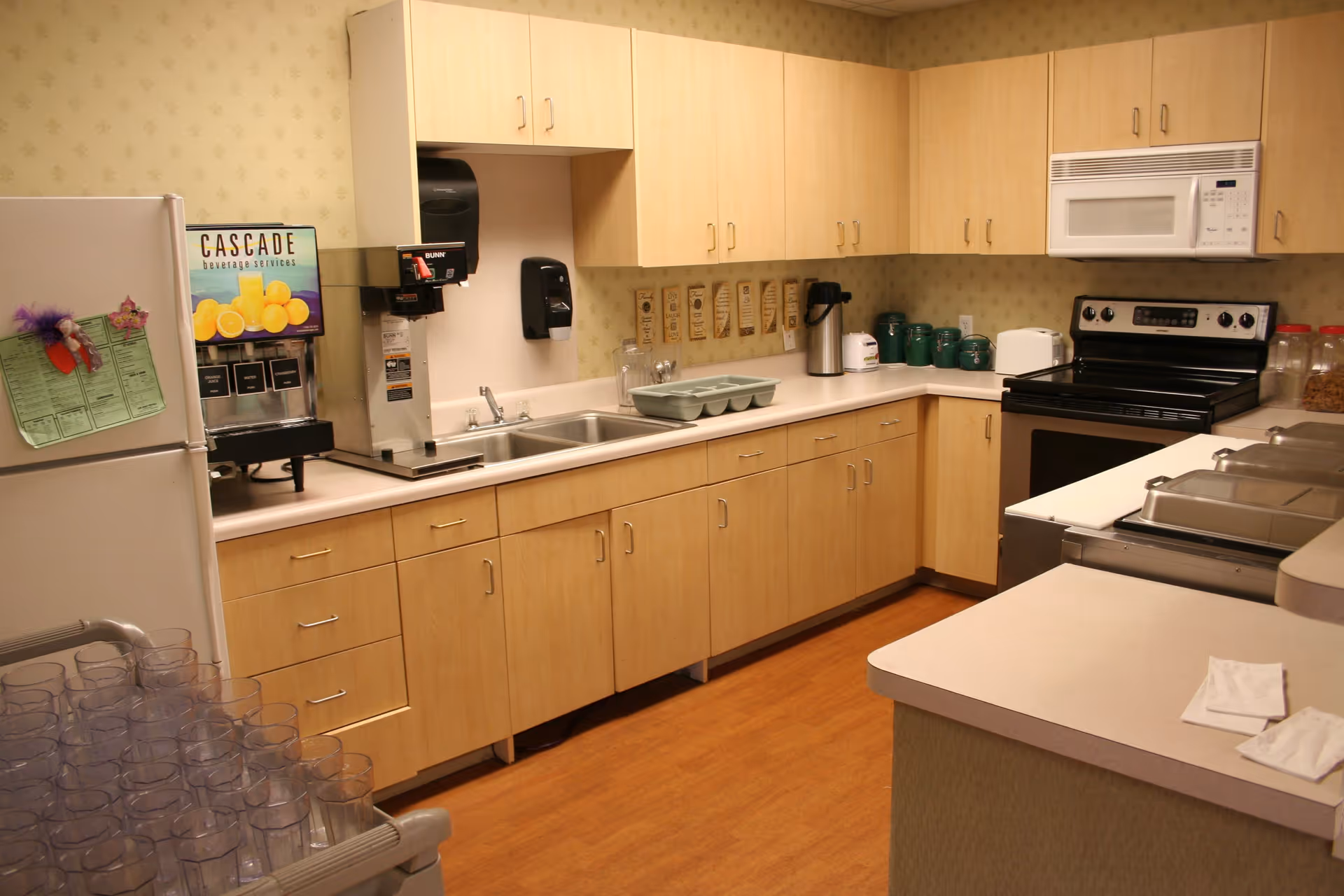 A clean kitchen area with light wood cabinets, a double sink, a white microwave above a black stove, a refrigerator, a beverage dispenser labeled 'Cascade Beverage Services', and various kitchen items including green canisters and a coffee dispenser.