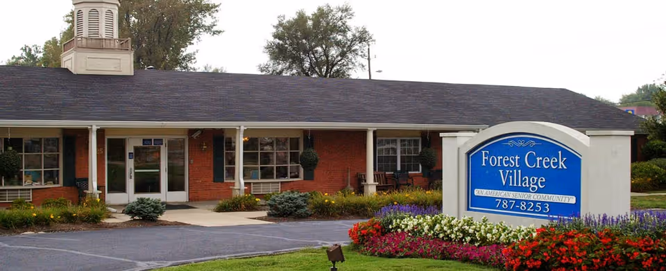 Front exterior of Forest Creek Village senior living building with the entrance and a large blue sign surrounded by flowerbeds.