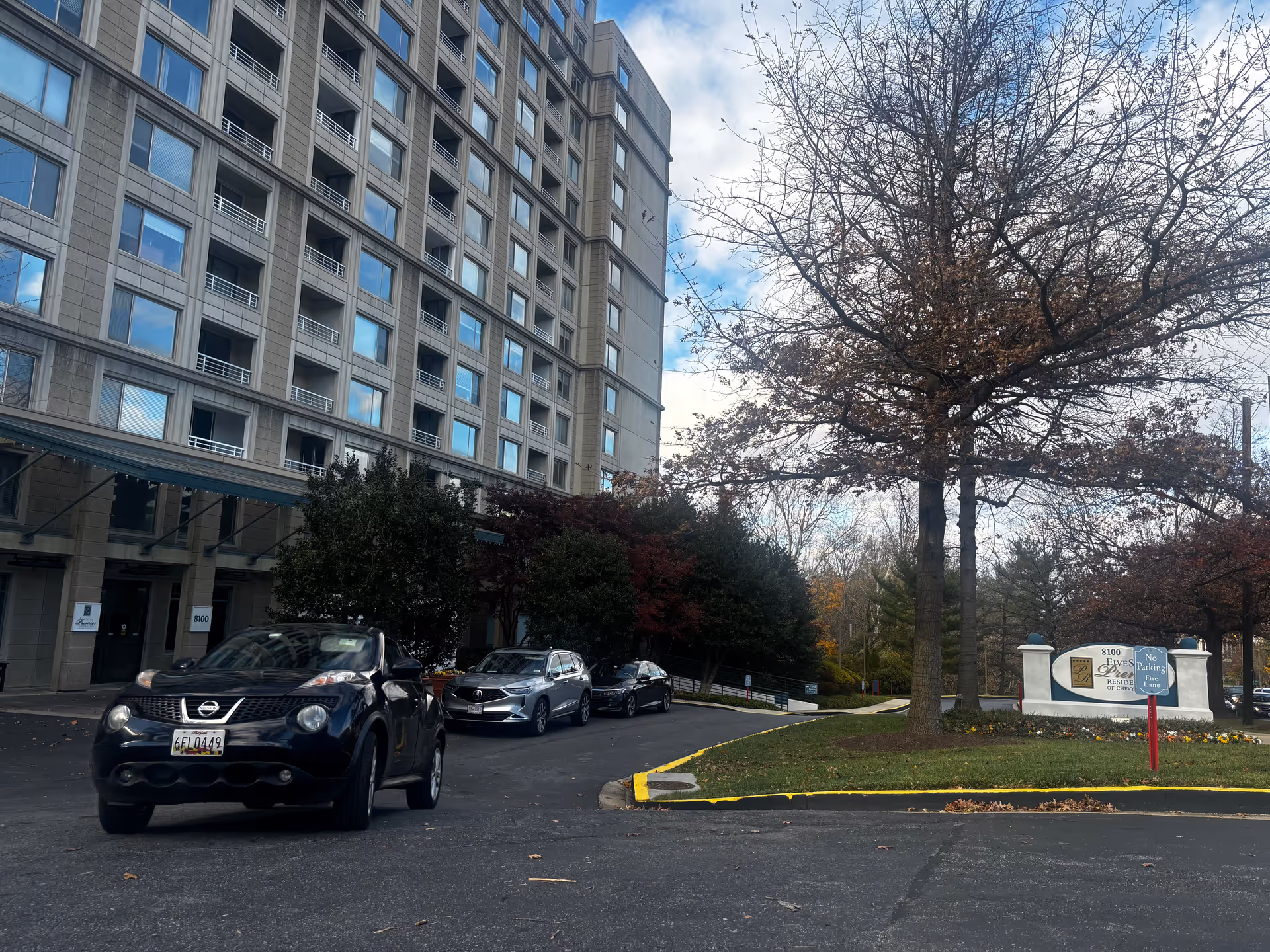 Exterior view of a multi-story residential building with several parked cars in front. There is a large tree with sparse leaves and a sign that reads 'Five Star Premier Residences of Chevy Chase' near the entrance driveway.