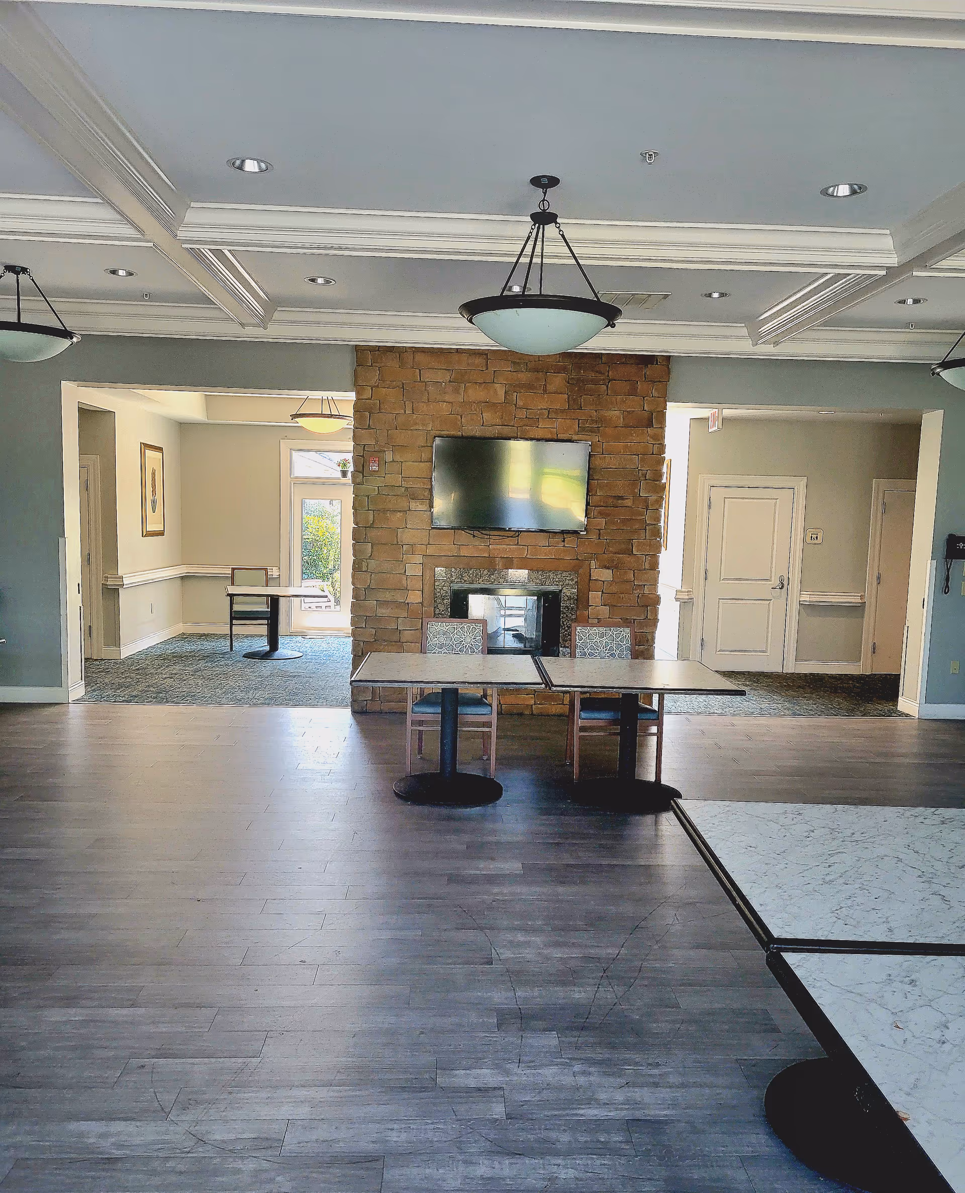 Interior view of a senior living facility common area with a stone fireplace and a mounted flat-screen TV. Two tables with chairs are placed in front of the fireplace. The room has wooden flooring, coffered ceiling with recessed lighting, and hanging light fixtures. Adjacent rooms with carpeted floors and additional tables are visible through open doorways.
