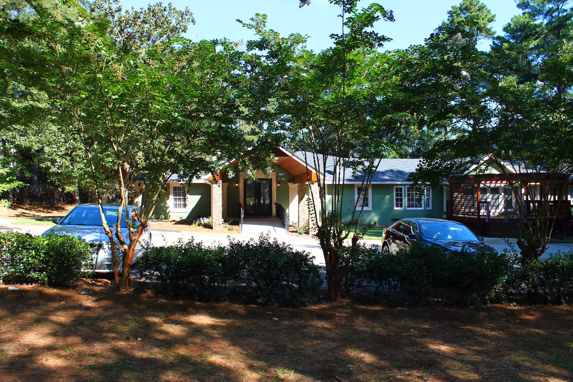Exterior view of a single-story green building with a covered entrance supported by stone pillars, surrounded by trees and bushes. Two cars are parked in front of the building under a clear blue sky.