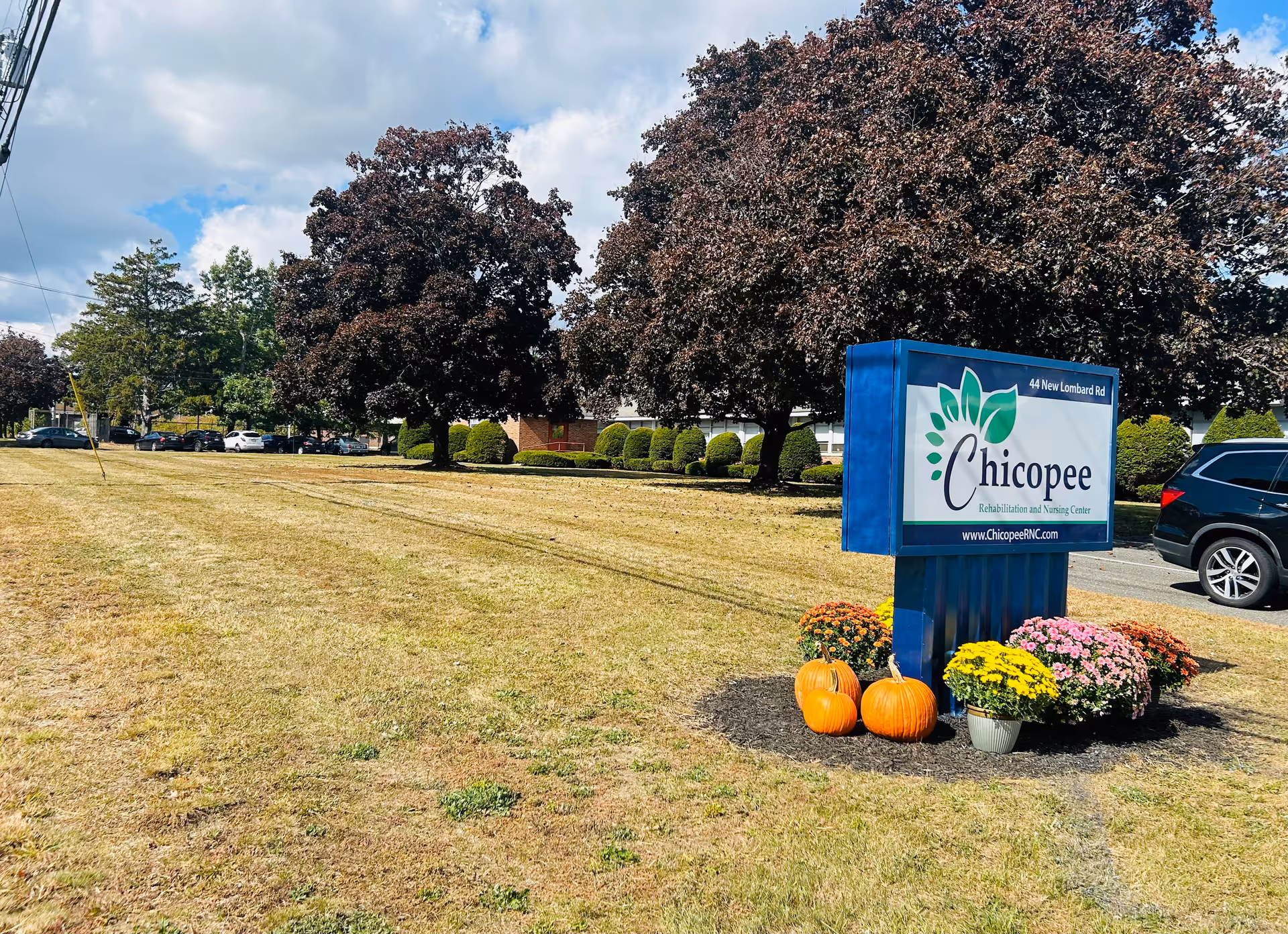 Blue sign reading 'Chicopee Rehabilitation and Nursing Center' on a grassy lawn decorated with pumpkins and flowers, with trees and the facility building in the background.