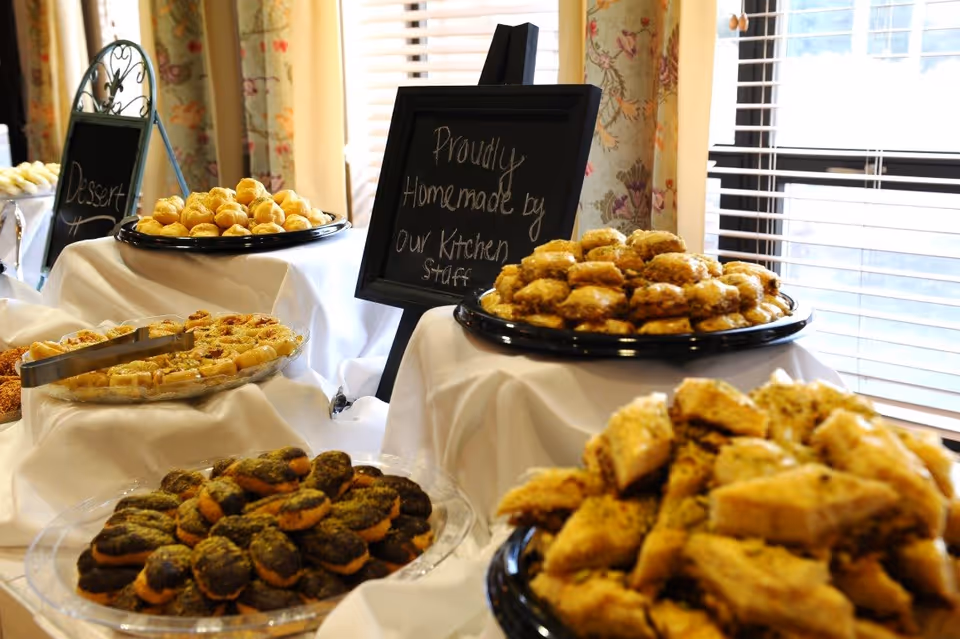 A buffet table with various trays of homemade baked goods and pastries displayed on white tablecloths near a window with floral curtains. Two small chalkboard signs are visible, one reading 'Proudly Homemade by our Kitchen staff' and the other partially showing the word 'Dessert'.