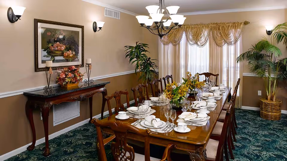 A formal dining room with a long wooden table set for a meal, including plates, cups, glasses, and napkins. The room features a green patterned carpet, beige walls with white trim, a chandelier, wall sconces, a framed painting, floral arrangements, and large windows with sheer curtains and gold drapes. There are also potted plants in the corners.