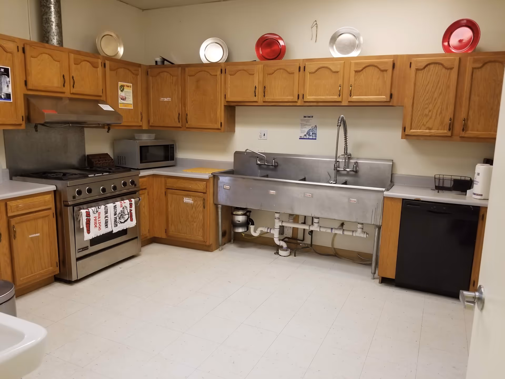 A kitchen with wooden cabinets, a stainless steel stove with a towel hanging on the handle, a microwave on the countertop, a large stainless steel triple sink with labeled sections, and a black dishwasher. Decorative plates are displayed on top of the cabinets.