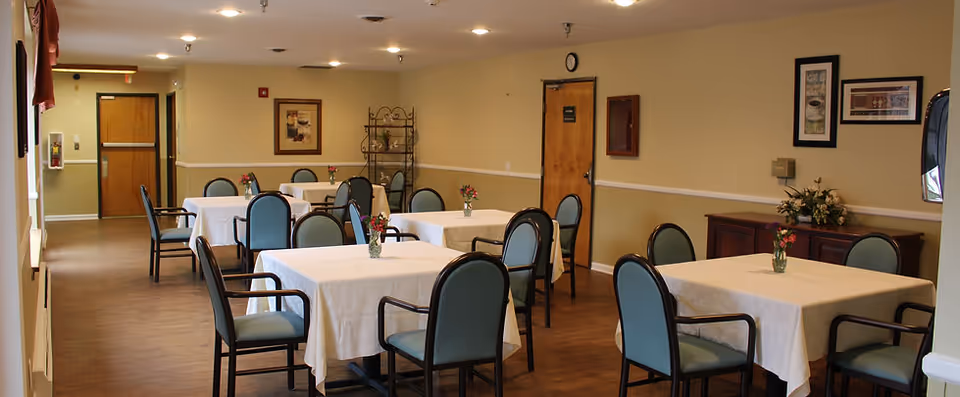 A dining room in Aspen Assisted Living with several square tables covered with white tablecloths, each table surrounded by blue cushioned chairs with black frames. Small flower vases are placed on each table. The room has beige walls with framed artwork, a wooden sideboard with a flower arrangement, and a wooden door. The floor is wooden, and the ceiling has recessed lighting.