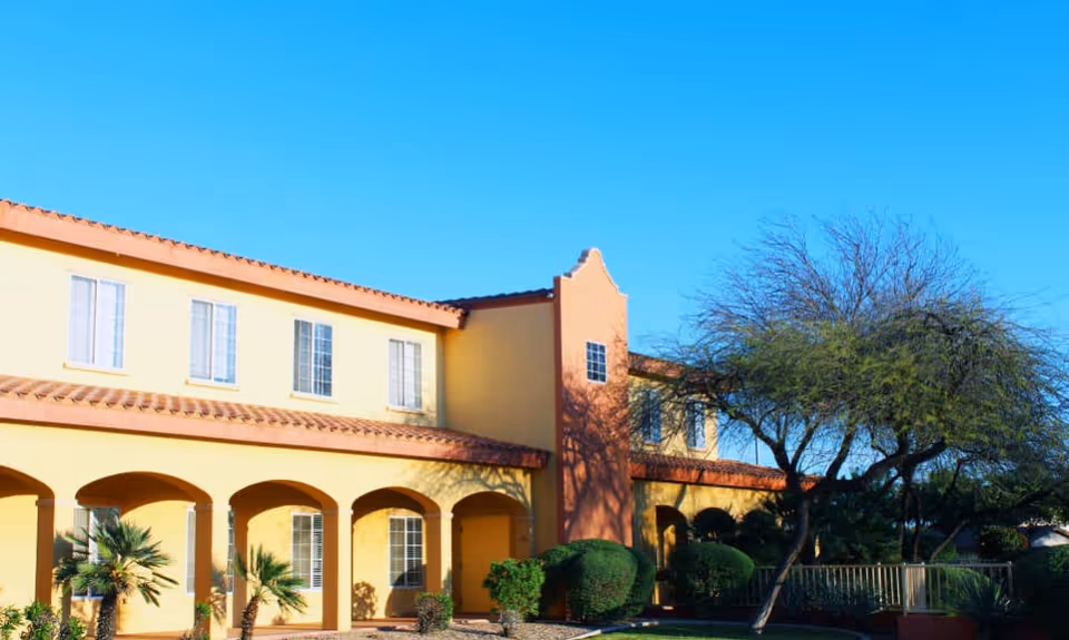 Exterior view of a two-story yellow building with a red-tiled roof, arched walkways, and several windows. There are small palm trees and bushes in front of the building, along with a larger tree on the right side under a clear blue sky.