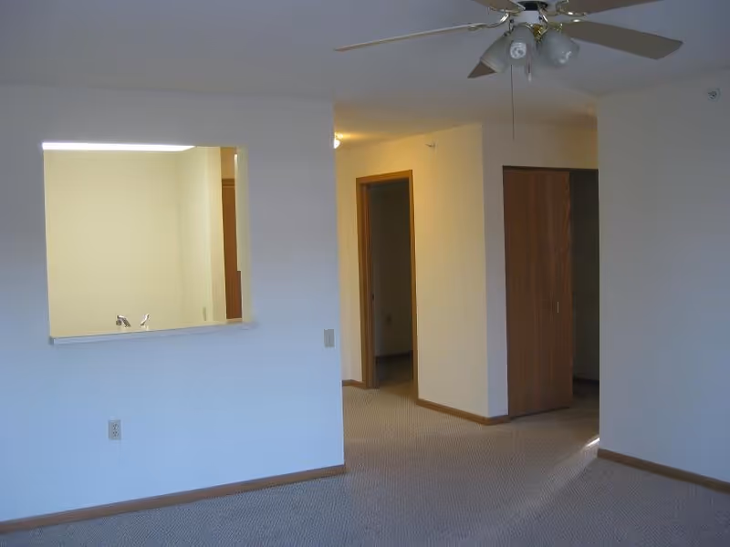Empty apartment interior with beige carpet, white walls, a ceiling fan with lights, a small pass-through window to the kitchen, and a hallway leading to other rooms with wooden door frames and closet doors.