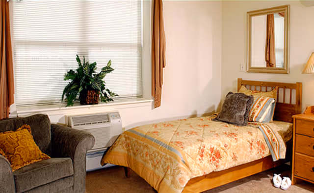 A cozy bedroom with a single wooden bed covered in a floral patterned quilt and multiple pillows. Next to the bed is a wooden nightstand with a lamp. A large window with blinds and brown curtains is behind a green armchair with a decorative pillow. A potted plant sits on the windowsill. A mirror hangs on the wall above the bed.
