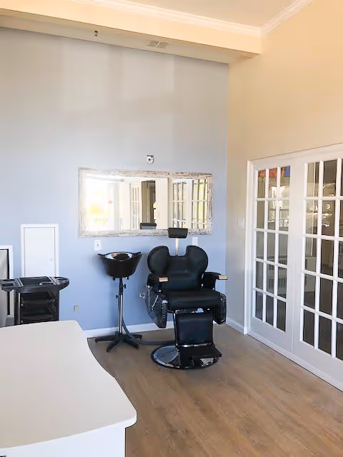 A small salon area inside a senior living facility with a black salon chair, a black hair washing basin, a large rectangular mirror on a light blue wall, and white French doors on the right side. The floor is wooden and the room is well-lit with natural light.