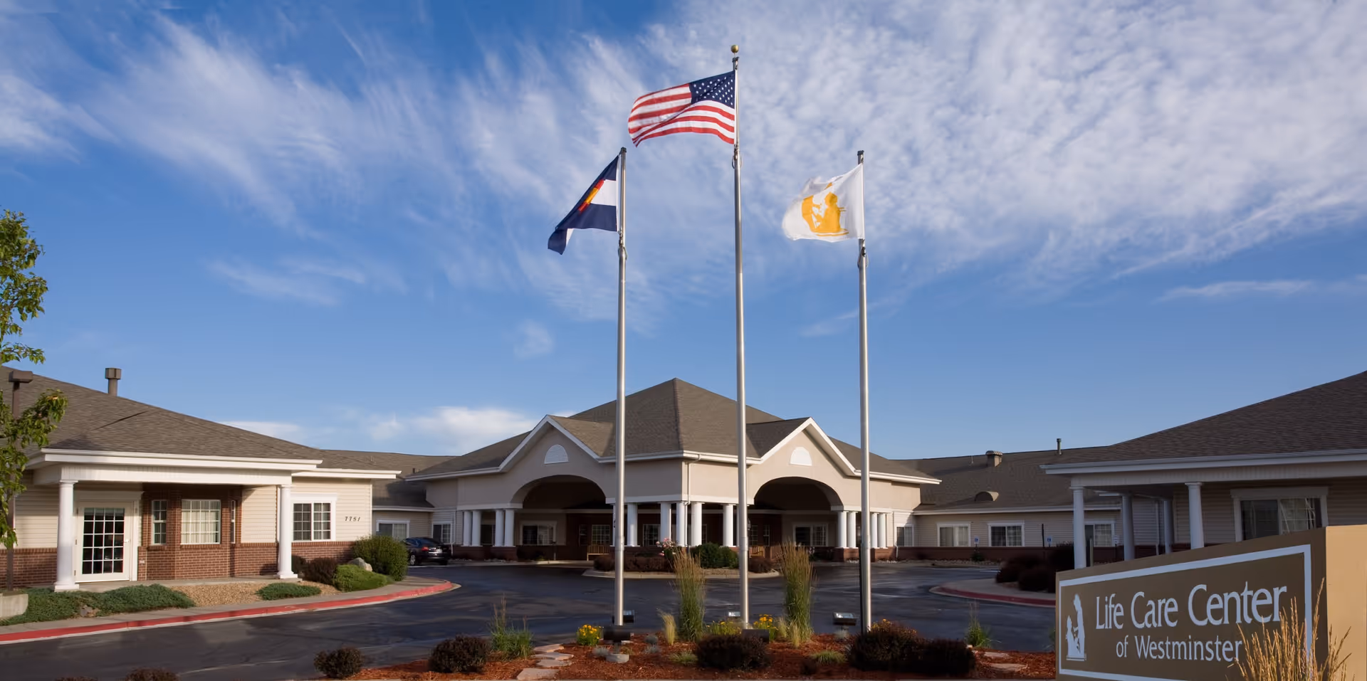 Front entrance of the Life Care Center of Westminster showing the building facade, three flagpoles, and landscaped driveway under a blue sky.