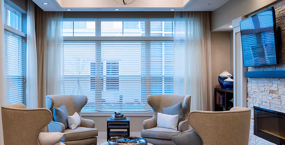 A cozy living room area with four beige armchairs arranged around a small round coffee table. Large windows with white blinds and sheer curtains let in natural light. A flat-screen TV is mounted above a stone fireplace on the right side, and a small bookshelf with decorative items is visible in the corner.