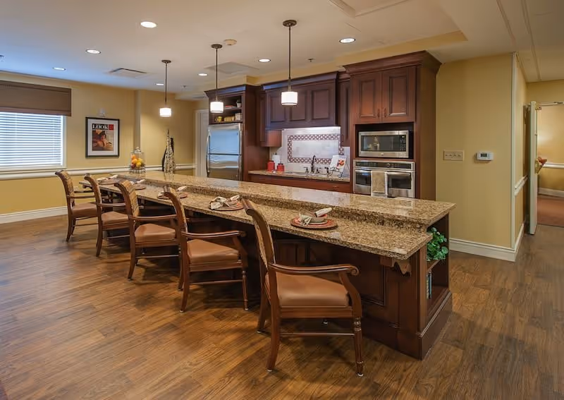 A warm communal kitchen featuring a long granite island with several wooden bar stools, pendant lights, and built-in stainless appliances.