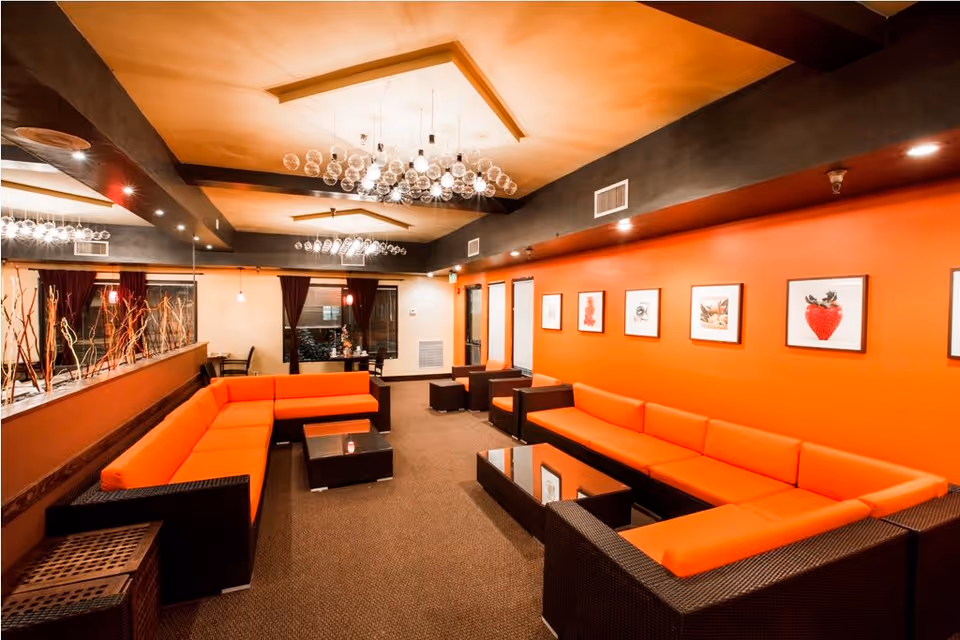 A modern senior living facility lounge with orange cushioned sectional sofas arranged around dark wood coffee tables. The room features an orange accent wall adorned with framed pictures of fruits, a beige carpeted floor, and a ceiling with contemporary light fixtures made of multiple glass bulbs. There is a glass partition with decorative branches on one side and a small dining area with chairs and tables in the background.